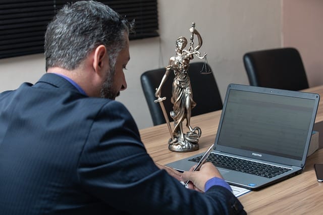 A man signing a document in front of a laptop. There is a statue of a lady with a sword in one hand and the scales of justice in the other on the table.